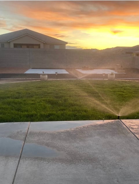 Residential backyard at sunset with sprinkler system active, concrete patio in foreground, and two-car garage with surrounding fenced area