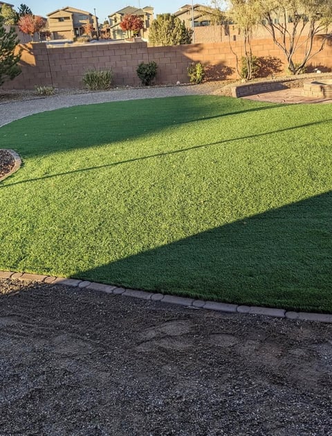 Desert backyard landscape with artificial green turf, gravel pathways, stucco wall, and two-story homes in background