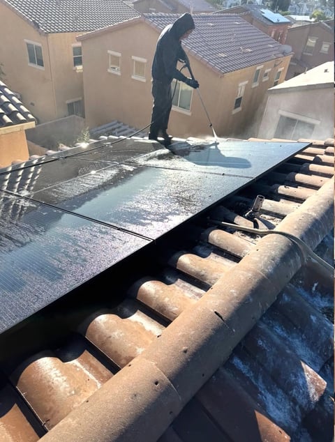 Worker in black protective gear applying coating to residential rooftop with terracotta tiles and adjacent homes visible below