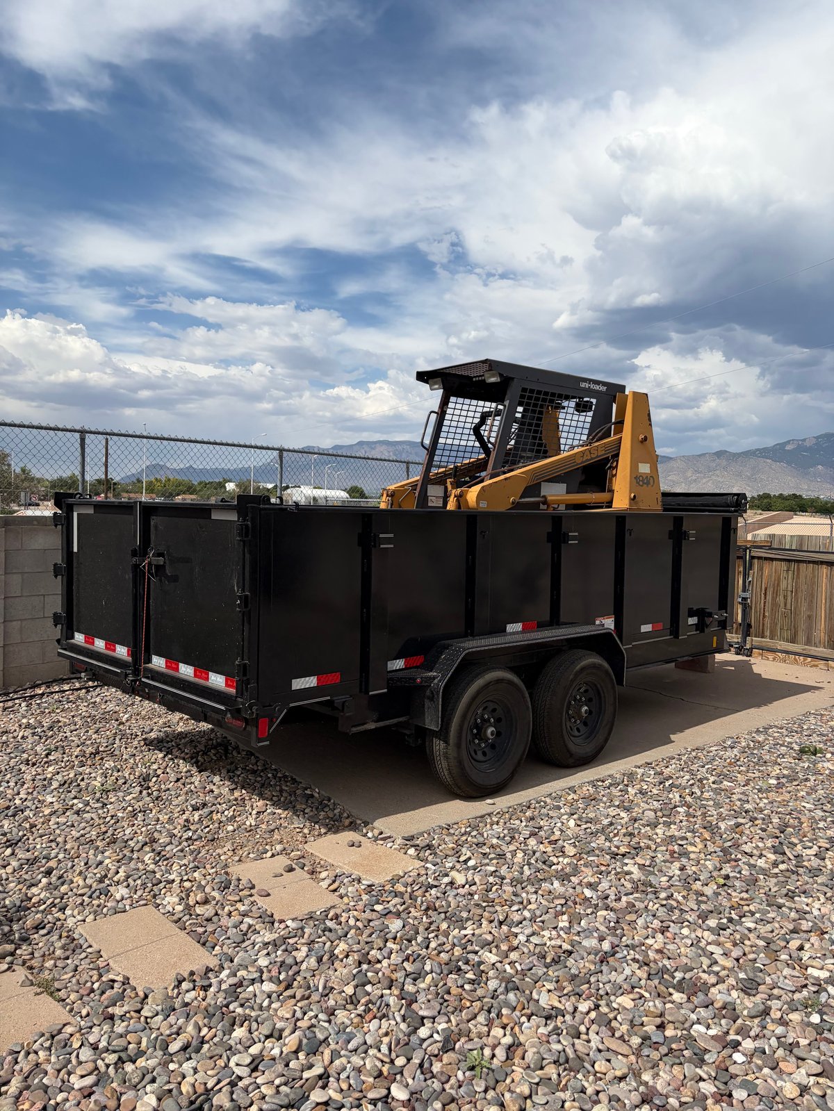 Yellow skid steer loader mounted on a black dual-axle dump trailer in a gravel lot
