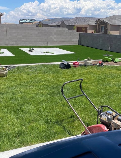 Lawn mower in foreground with lush green residential backyard, concrete block walls, and suburban homes under blue sky with clouds
