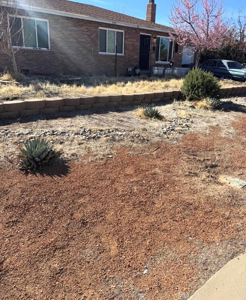 Single-story brick house with desert landscaping, sparse vegetation, gravel yard, and parked vehicle