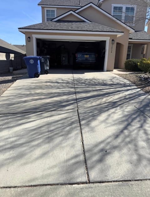 Two-car garage with open door showing parked vehicles, concrete driveway with tree shadows, tan stucco house exterior, clear blue sky